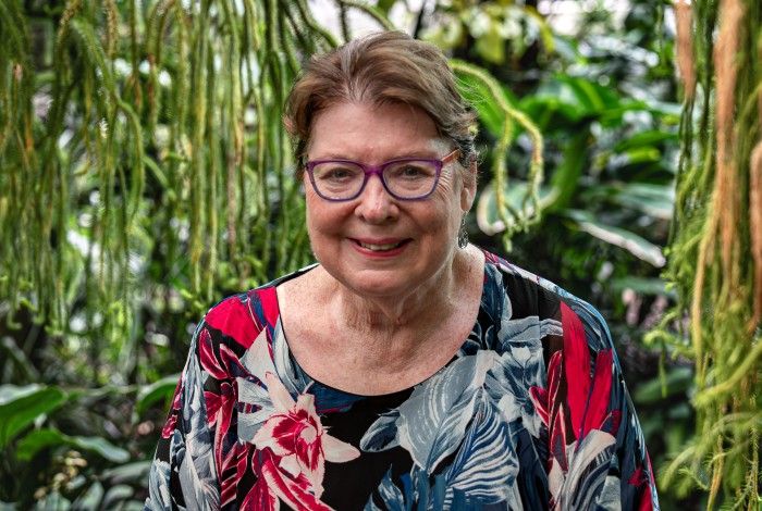 An older woman with short brown hair and purple glasses smiles warmly at the camera. She is wearing a colorful floral top with red, white, and blue patterns. Lush green tropical plants hang in the background, creating a vibrant, natural setting.