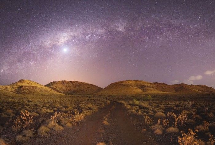 A night time landscape in the Pilbara region, showing a rugged dirt road leading through low, rocky hills under a star-filled sky. The Milky Way arcs clearly across the sky, with a bright planet or star shining near the centre. The foreground is covered in sparse, dry vegetation and small shrubs, typical of the Australian outback.
